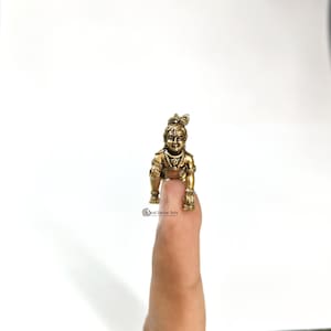 May include: A small, brass-colored statue of a deity in a crawling pose. The figure is adorned with a crown and jewelry. The statue is held on a finger, with the logo "Desi Decor Arts" visible at the base.