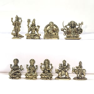 Könnte beinhalten: Eine Sammlung kleiner, goldfarbener Statuen verschiedener Gottheiten, darunter Figuren mit mehreren Armen und sitzenden Formen. Die Statuen sind auf einer weißen Oberfläche angeordnet und zeigen komplizierte Details und traditionelle Posen. Die Sammlung enthält eine Pfauenstatue.