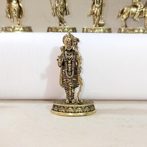 May include: A gold-colored metal figurine of a Hindu deity holding a bow and arrow. The figurine is standing on a decorative base.