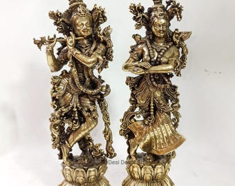 Puede incluir: Dos estatuas doradas y ornamentadas de deidades. Una figura toca la flauta, la otra tiene los brazos cruzados. Ambas se alzan sobre bases de flores de loto, con detalles intrincados en su vestimenta y tocados. Para decoración o fines religiosos.