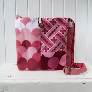 May include: A pink and red patchwork tote bag with a geometric pattern and a brown button. The bag has a long strap and is made of fabric.