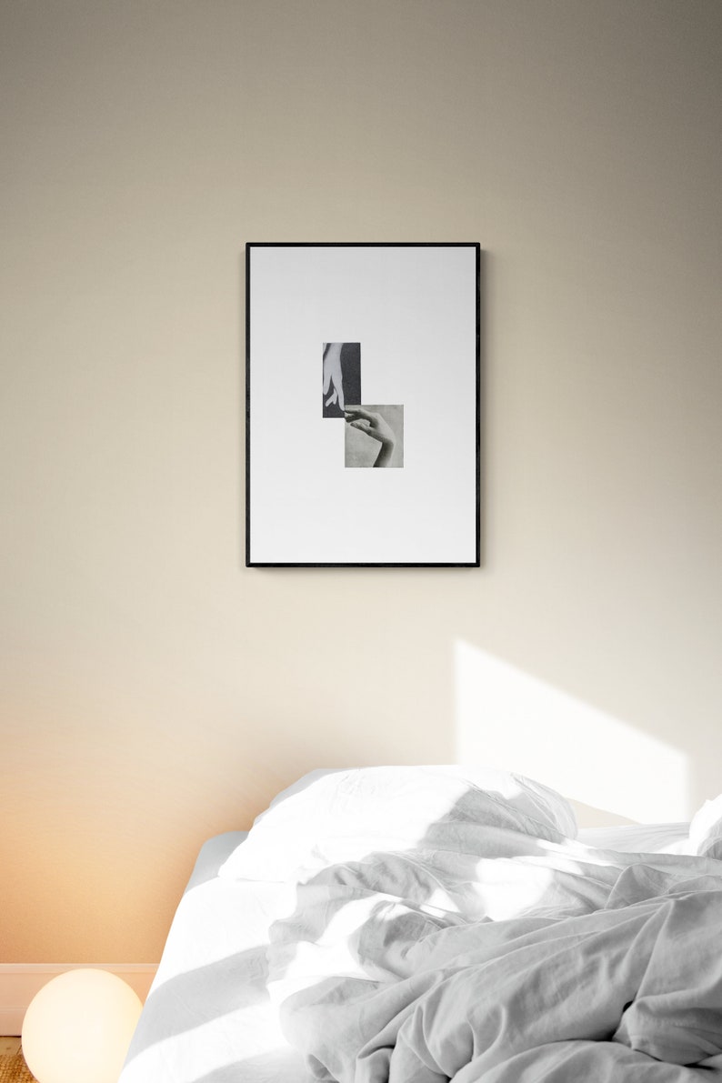 May include: A black framed picture with a white background and two black and white photographs of hands. The picture is hanging on a beige wall above a white bed with a white duvet and pillows.