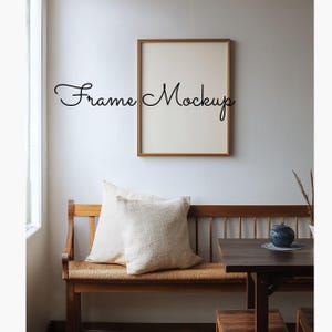 Puede incluir: Una maqueta enmarcada con el texto "Frame Mockup" en una fuente cursiva se muestra en una pared blanca. Debajo, se ve un banco de madera con dos almohadas de color crema. Una mesa de madera oscura y un jarrón de cerámica azul también están en la escena.