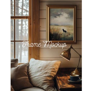 Op de afbeelding: Een ingelijst landschapsschilderij met een gouden lijst hangt aan een wand met houten panelen. Het schilderij toont een veld met een donker dierensilhouet. Een messing bureaulamp en een kopje thee staan op een houten tafel. De tekst "Frame Mockup" staat op de afbeelding.