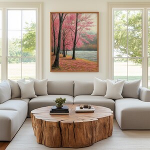 May include: A framed landscape painting depicts pink flowering trees along a riverbank. The artwork hangs above a light gray sectional sofa with white pillows. A large, round wooden coffee table sits on a neutral rug.