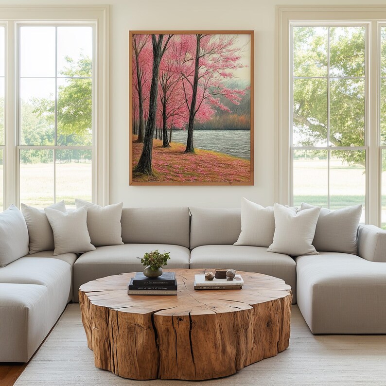May include: A framed landscape painting depicts pink flowering trees along a riverbank. The artwork hangs above a light gray sectional sofa with white pillows. A large, round wooden coffee table sits on a neutral rug.