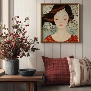 May include: A framed illustration of a woman with long brown hair, closed eyes, and a red dress. The woman is surrounded by white flowers and green leaves. The frame is gold and the background is a white wood panel.