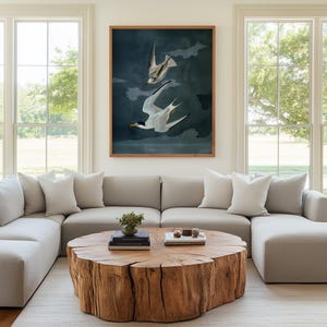 May include: A living room featuring a large, light gray sectional sofa, white pillows, and a round wooden coffee table. A framed artwork of two birds in flight hangs above the sofa. Large windows provide natural light.