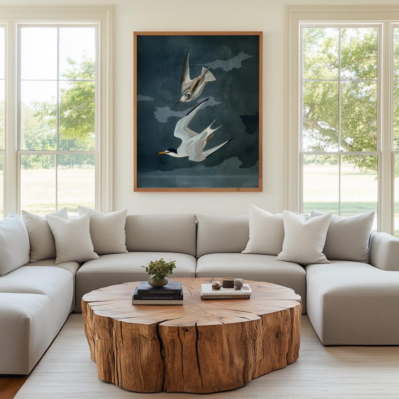 May include: A living room featuring a large, light gray sectional sofa, white pillows, and a round wooden coffee table. A framed artwork of two birds in flight hangs above the sofa. Large windows provide natural light.