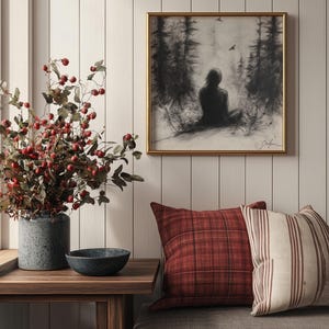 May include: A gray couch with red and white patterned throw pillows. A framed black and white drawing of a person sitting in a forest hangs on the wall above the couch. A vase with red berries and green leaves sits on a wooden table in front of the couch.