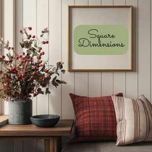 May include: A framed print with a green background and the text "Square Dimensions" in black cursive writing. The print is hanging on a white wood panel wall. A vase with red berries and green leaves sits on a wooden table in front of the print. Two pillows, one red plaid and one white with brown stripes, are on a couch in the background.