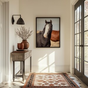 May include: A black and white horse with a white blaze on its face stands in a field, looking directly at the camera. The horse is framed in a black picture frame and hangs on a white wall. A wooden console table with a brown top and black legs sits in front of the picture. A vase with dried branches sits on the table.