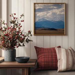 May include: A living room with a wooden bench, two pillows, a small side table, a vase with red berries, and a framed picture of a mountain range with snow-capped peaks.