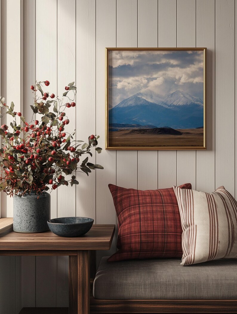 May include: A living room with a wooden bench, two pillows, a small side table, a vase with red berries, and a framed picture of a mountain range with snow-capped peaks.