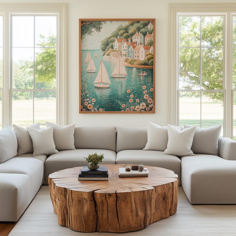 May include: A framed watercolor painting depicts sailboats on a turquoise sea, with a coastal village in the background. The artwork hangs above a light gray sectional sofa, complemented by a natural wood coffee table and white pillows. The room has large windows.