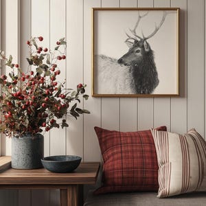 May include: A framed black and white print of a deer with antlers, hanging on a white wood paneled wall. A vase with red berries and a bowl are on a wooden table in front of a gray couch with red and white patterned pillows.