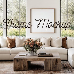 Peut inclure: Intérieur de salon avec un grand canapé blanc, des coussins décoratifs et une table basse en bois. Une toile vierge encadrée est accrochée au mur, avec le texte "Frame Mockup" en écriture cursive. Un vase de fleurs est posé sur la table.