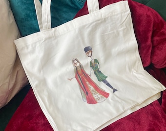 May include: A white tote bag with a printed design of a couple in traditional clothing. The woman is wearing a red and gold dress and the man is wearing a green and gold outfit.