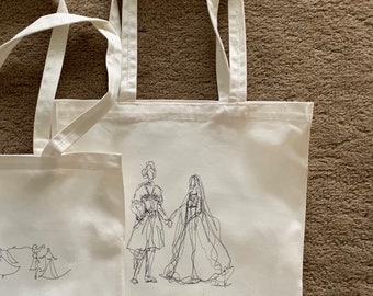 May include: Two white canvas tote bags with black line drawings of people. One bag features a couple holding hands, the other bag features a group of people dancing. The bag with the couple has the text "Wedding Day" written below the drawing.
