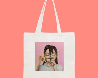 May include: A white tote bag with a pink background and a painting of a person cutting their hair with yellow scissors. The person has brown hair and is wearing a light grey shirt.