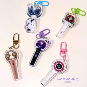 May include: Five different keychains featuring various K-pop groups. The keychains are made of clear acrylic with glitter and colorful designs. The keychains feature the logos of BTS, Twice, and other K-pop groups.