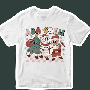 May include: White t-shirt featuring a retro-style graphic of a Christmas tree, snowman, and Santa Claus with the text "ABA CREW". The characters are holding a data chart and candy cane. The design has a vintage, cartoonish aesthetic.