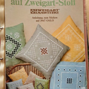 Può includere: Una copertina di libro con il titolo "Schöner Sticken auf Zweigart-Stoff" e il logo Zweigart & Sawitzki. La copertina presenta una varietà di cuscini ricamati colorati con diversi motivi.