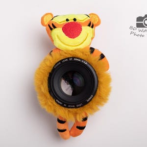 May include: A plush tiger-themed camera lens cover with a Canon lens. The cover is orange and yellow with black stripes and a red nose. The lens is black with white text that reads "CANON LENS EF 50mm 1:1.4".