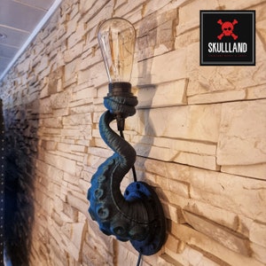 Könnte beinhalten: Eine Wandleuchte in Form eines Krakenarmes mit einer Glühbirne im Vintage-Stil. Der Arm ist in einem dunklen Teal-Ton lackiert und hat eine schwarze Metallbasis. Die Leuchte ist an einer Backsteinwand montiert.