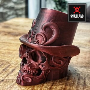 Könnte beinhalten: Ein 3D-gedruckter Schädel, der einen Zylinder mit komplizierten Details trägt. Der Schädel und der Hut sind tiefrot. Der Schädel hat eine Steampunk-Ästhetik mit Zahnrädern und anderen mechanischen Elementen.