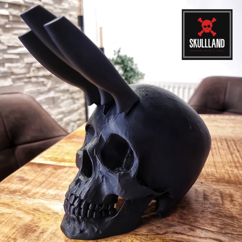 May include: A black skull-shaped knife holder with three black knives sticking out of the top. The skull is sitting on a wooden surface. The text "SKULLLAND" is visible in the upper right corner of the image.