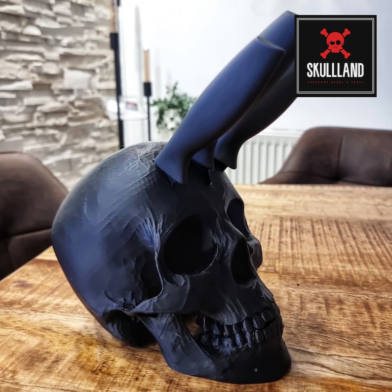 May include: A black skull sculpture with two black knives sticking out of the top of the skull. The skull is sitting on a wooden surface. The text "SKULLLAND" is visible in the background.