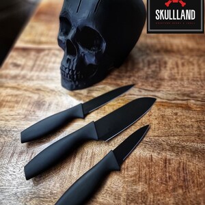 May include: Three black kitchen knives with black handles and a black skull shaped knife holder on a wooden surface. The skull has three holes in the top and the text "SKULLLAND" is on a black and red square.
