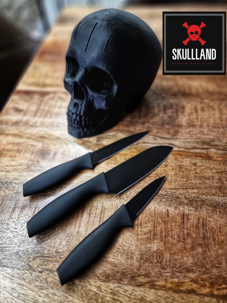 May include: Three black kitchen knives with black handles and a black skull shaped knife holder on a wooden surface. The skull has three holes in the top and the text "SKULLLAND" is on a black and red square.