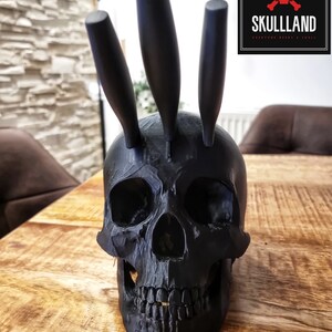 May include: A black skull-shaped knife holder with three black knives sticking out of the top. The skull is sitting on a wooden surface.