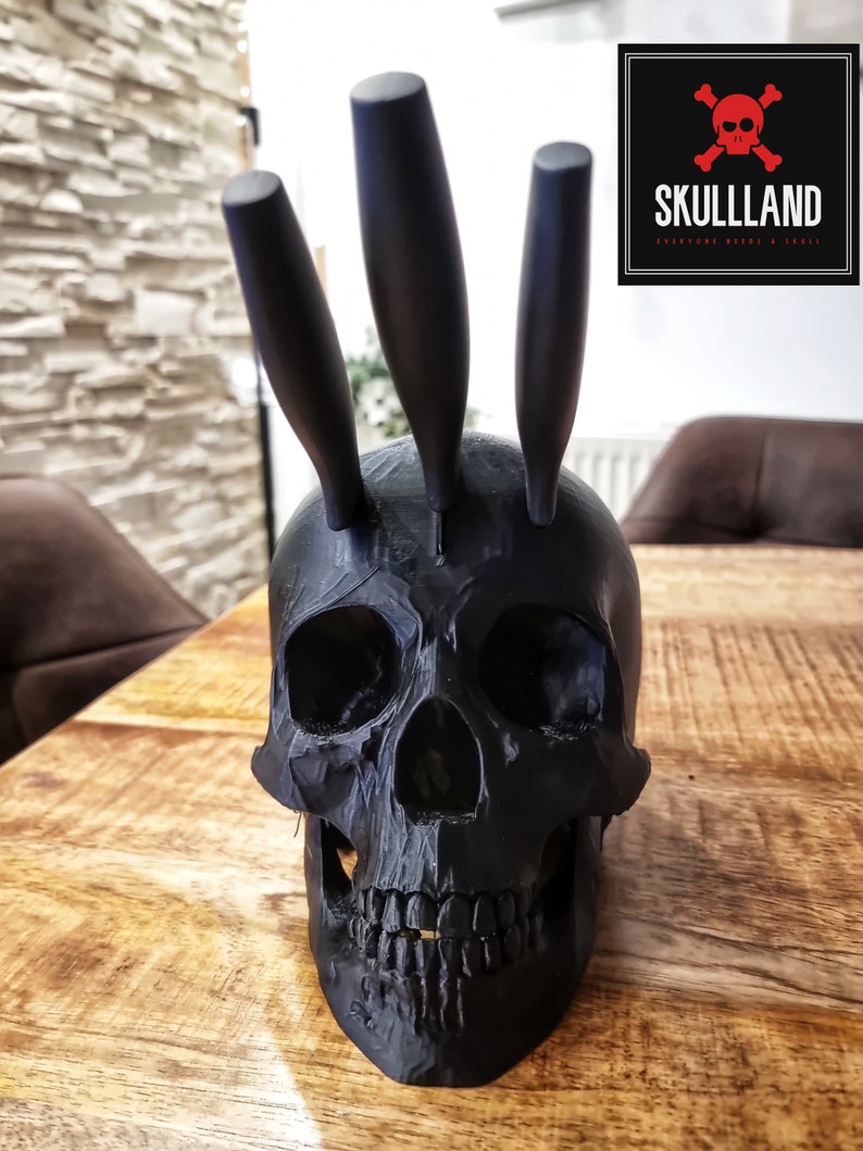 May include: A black skull-shaped knife holder with three black knives sticking out of the top. The skull is sitting on a wooden surface.