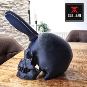 May include: A black skull-shaped knife holder with two black knives sticking out of the top. The skull is sitting on a wooden table. The text "SKULLLAND" is visible in the upper right corner of the image.