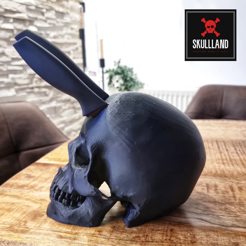 May include: A black skull-shaped knife holder with two black knives sticking out of the top. The skull is sitting on a wooden table. The text "SKULLLAND" is visible in the upper right corner of the image.