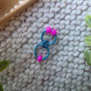 Pu&ograve; includere: Un paio di orecchini a cerchio blu con perline quadrate rosa acceso. Gli orecchini sono collegati e poggiano su una superficie strutturata bianco sporco. Gli orecchini creano un vivace contrasto con lo sfondo neutro.