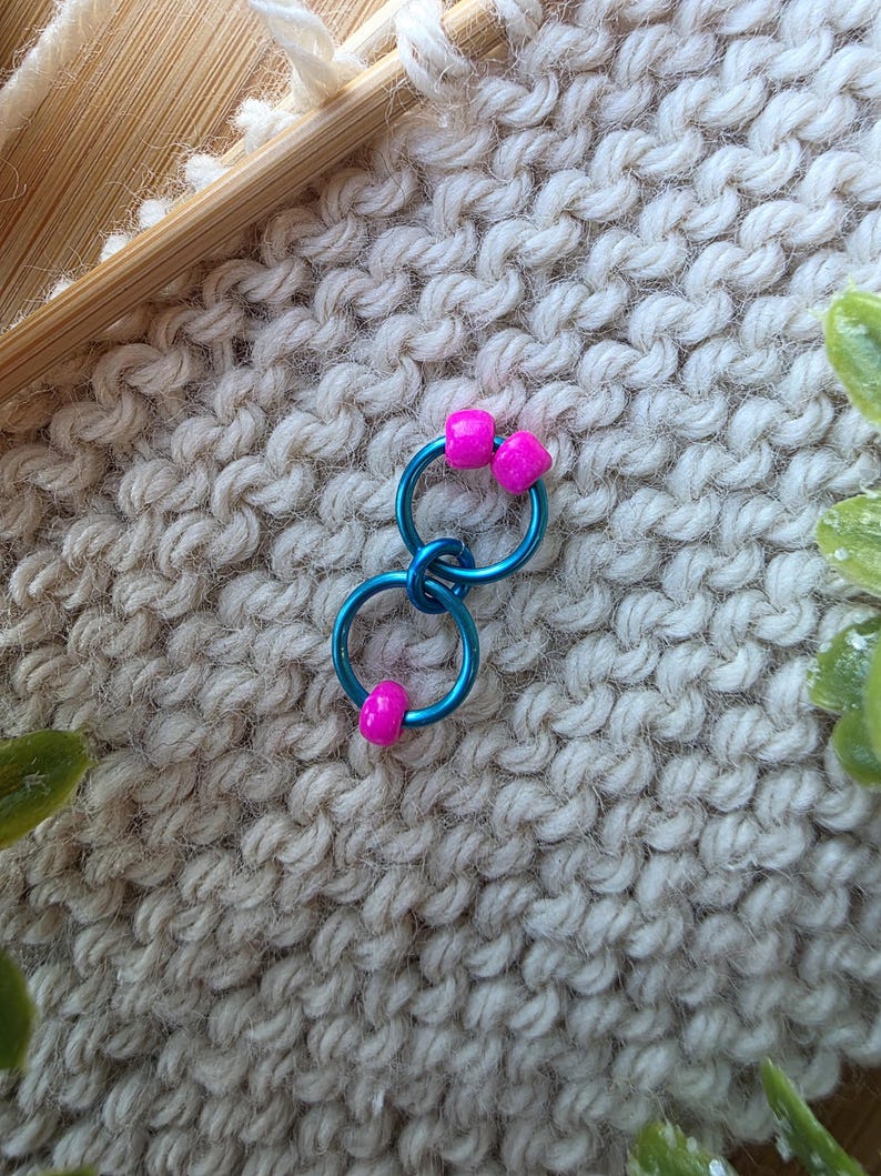 Pu&ograve; includere: Un paio di orecchini a cerchio blu con perline quadrate rosa acceso. Gli orecchini sono collegati e poggiano su una superficie strutturata bianco sporco. Gli orecchini creano un vivace contrasto con lo sfondo neutro.