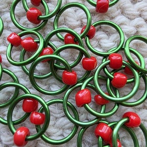 May include: A collection of stitch markers, each consisting of a smooth, circular green metal ring adorned with one or two small, glossy red beads. These functional knitting accessories are arranged in a scattered pattern, showcasing the vibrant contrast between the metallic green hoops and the bright red spherical beads.