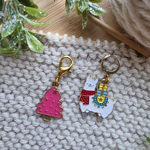 May include: Two gold-colored charms on a light beige knitted background. One charm is a pink Christmas tree, and the other is a white llama wearing a red scarf and carrying a blue and yellow pack. The charms are likely for a bracelet or necklace.