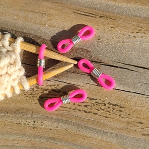May include: Four pink silicone stitch markers with silver metal rings. The markers are on a wooden surface.