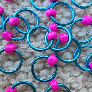 Pu&ograve; includere: Primo piano di diversi anelli metallici color teal, ciascuno adornato con una vivace perla rosa. Gli anelli sono disposti in un motivo sparso, creando un display visivamente accattivante. Lo sfondo &egrave; una superficie strutturata bianco sporco.