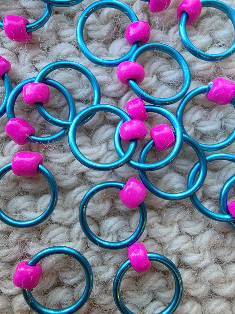 Pu&ograve; includere: Primo piano di diversi anelli metallici color teal, ciascuno adornato con una vivace perla rosa. Gli anelli sono disposti in un motivo sparso, creando un display visivamente accattivante. Lo sfondo &egrave; una superficie strutturata bianco sporco.