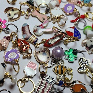 May include: A collection of colorful, decorative charms with gold-tone rings. The charms feature various designs, including a fried egg, a red shoe, a cupcake, a bee, a flag, and a rabbit. The charms are likely for jewelry or keychains.