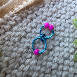 Pu&ograve; includere: Primo piano di un progetto di lavoro a maglia in corso, con segnapunti blu e perline rosa. Il progetto utilizza ferri da maglia in legno e filato bianco sporco, con i segnapunti attaccati al tessuto lavorato a maglia.