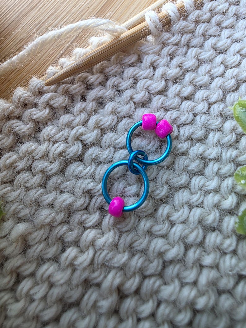 Pu&ograve; includere: Primo piano di un progetto di lavoro a maglia in corso, con segnapunti blu e perline rosa. Il progetto utilizza ferri da maglia in legno e filato bianco sporco, con i segnapunti attaccati al tessuto lavorato a maglia.