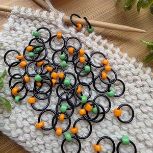 May include: A collection of black metal stitch markers, each featuring a single small bead in either bright orange or light green. These circular rings are designed for knitting projects and are scattered across a piece of off-white knitted fabric.