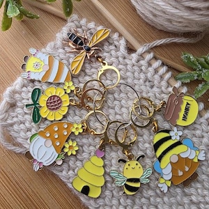 May include: A collection of colorful bee-themed stitch markers. The markers include a bee, a gnome, a beehive, and a honey pot. The markers are attached to gold-colored rings and are set on a knitted background.